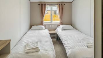 A room with two single beds, a window with curtains, a wooden bench, and a view of a yellow building outside.