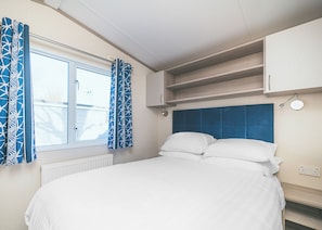 Une chambre à coucher comprenant un lit, une fenêtre, des rideaux, une tête de lit, une étagère et un luminaire fixé au mur.