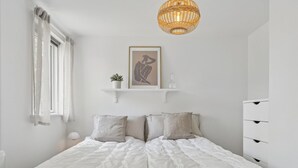 Un dormitorio con una cama grande, una mesita de noche, una lámpara colgante, un ventanal con cortinas y una estantería con un cuadro enmarcado y una planta pequeña.