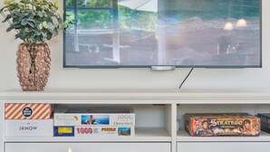 Una sala de estar con un televisor montado en la pared, una planta en maceta y una mesa con una vela. Hay cajas de rompecabezas en el estante inferior.