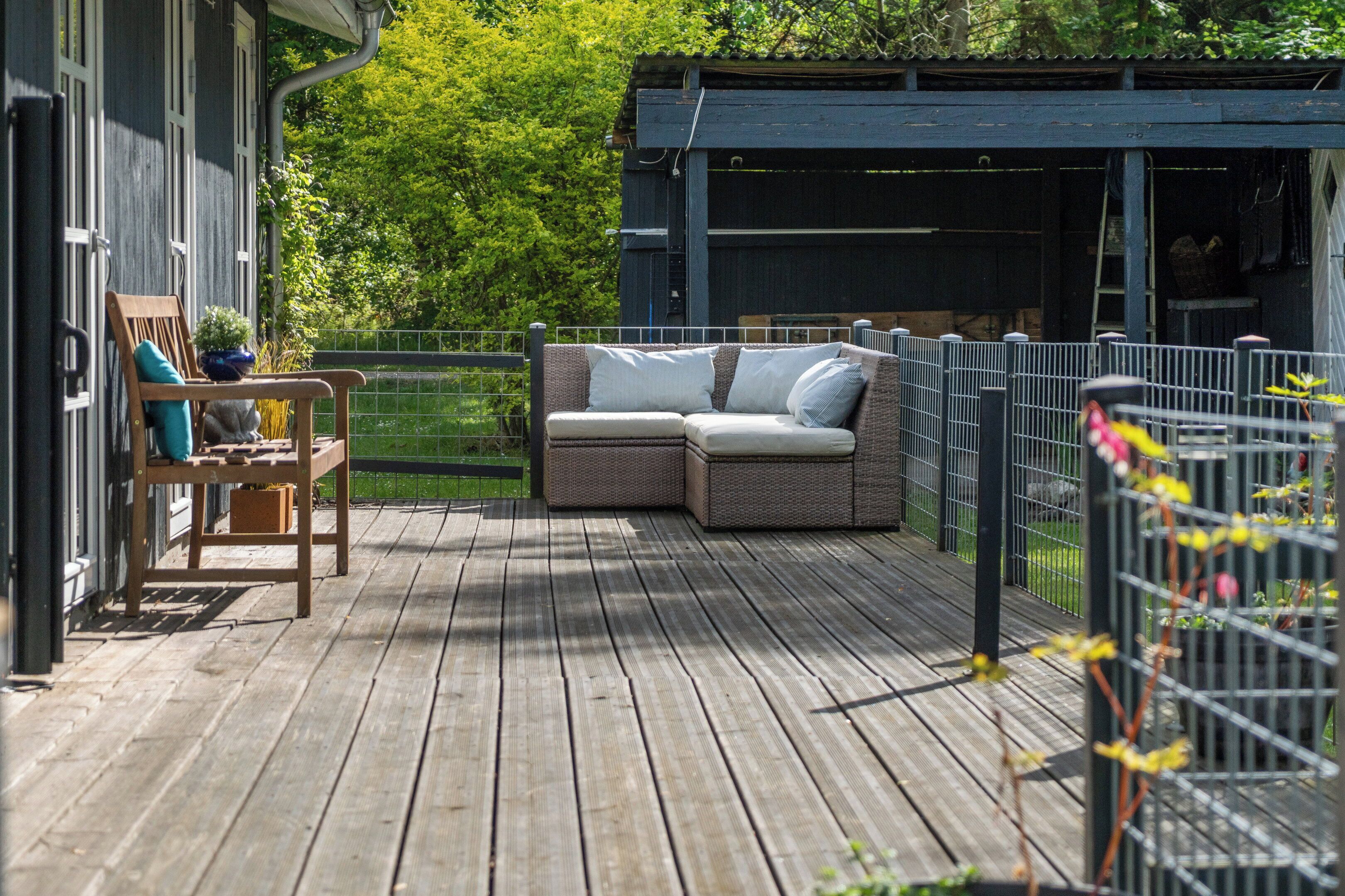 A wooden deck with a wicker sofa and a wooden chair, a small table with a plant, and a metal fence.