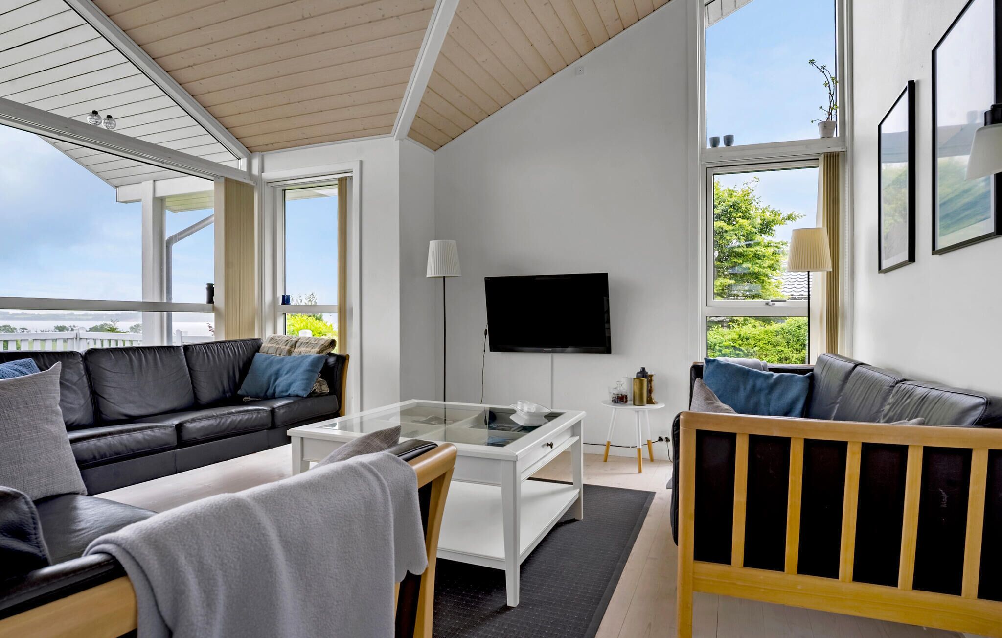 A modern living room with a black leather sofa, a white coffee table, and a wooden bench. A flat-screen TV is mounted on the wall, and there are large windows providing a view of the outdoors.