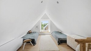 Ein kleines, dreieckiges Schlafzimmer im Dachgeschoss mit zwei Betten, einer hölzernen Bank und Blick auf ein Haus durch ein Fenster.