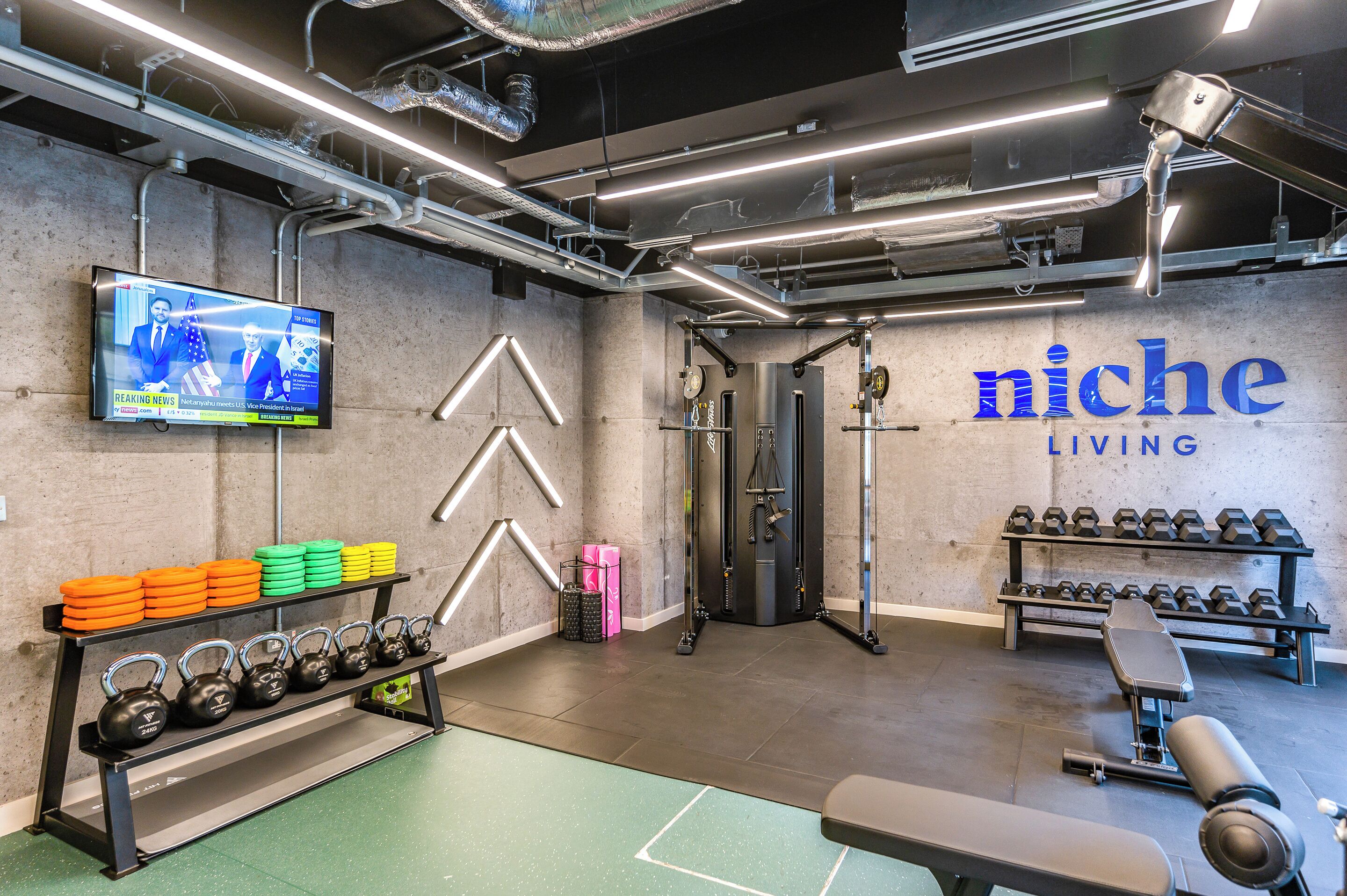 Una palestra con attrezzature per l'allenamento con i pesi, un televisore che mostra le notizie e un muro con la scritta 'niche LIVING'.