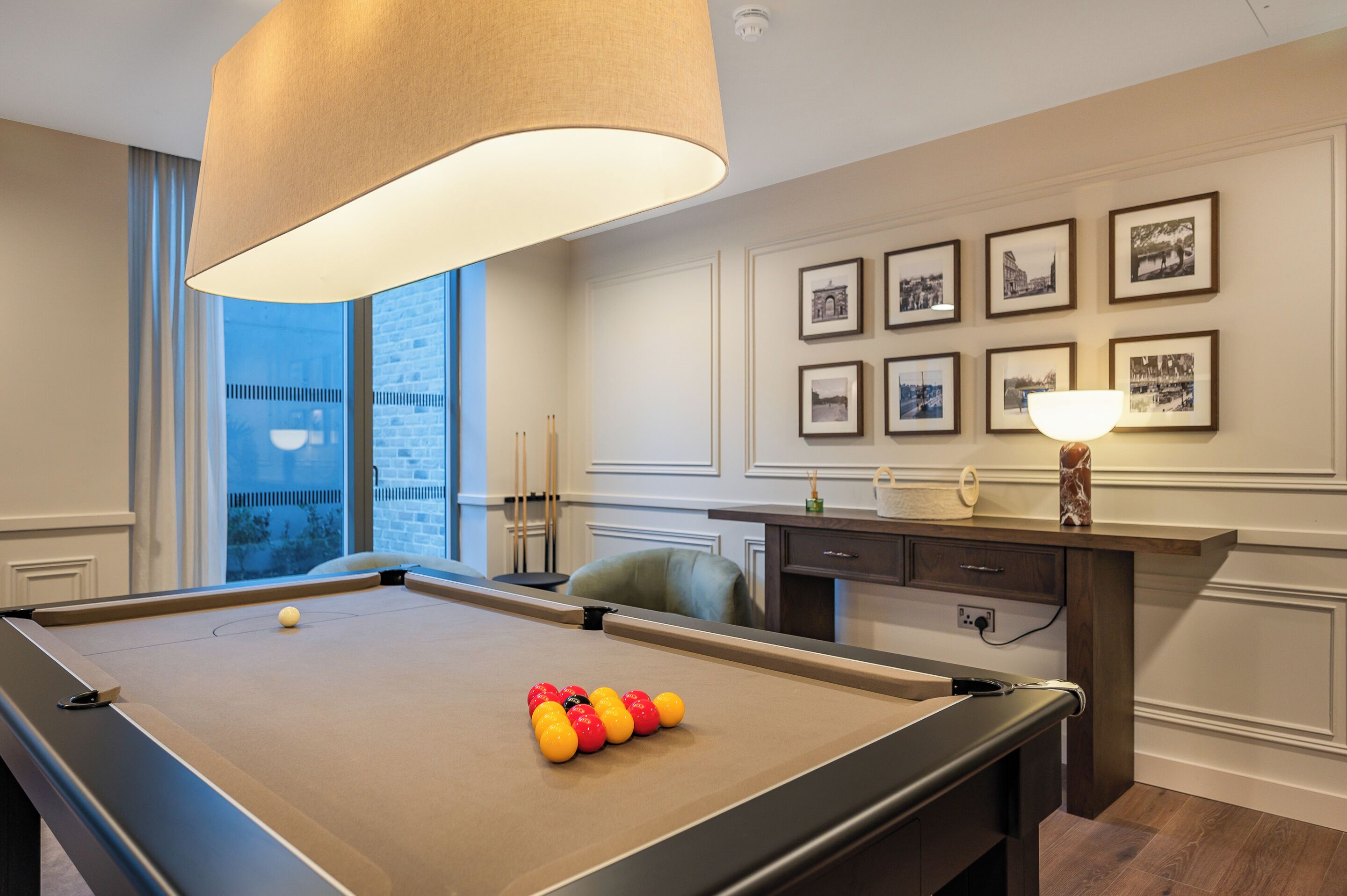 A pool table with balls, a lamp, framed pictures on the wall, and a view of a building outside.