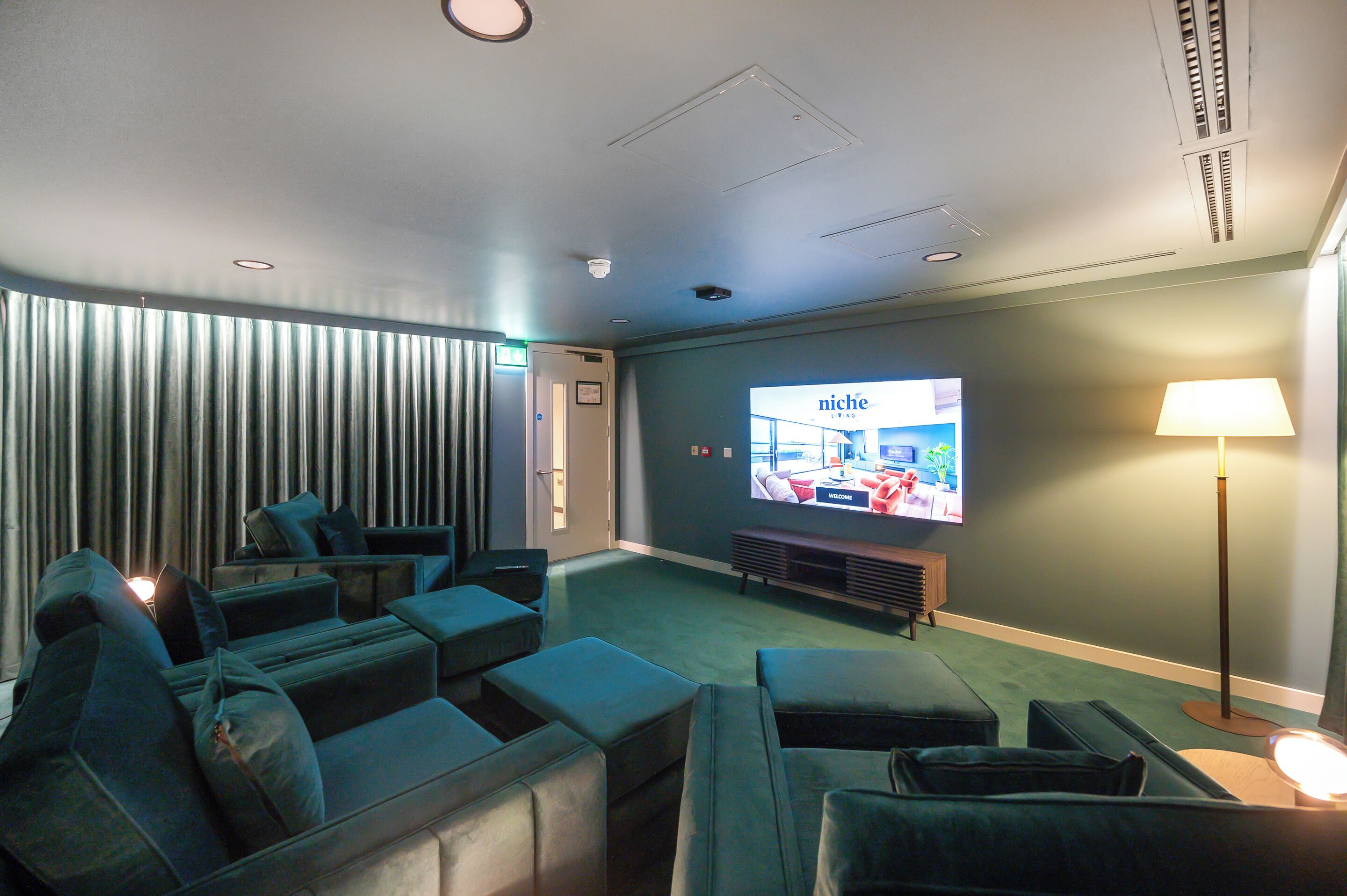 Un salón moderno con televisor de pantalla plana mostrando 'Niche Living', una zona de sofás y una lámpara.
