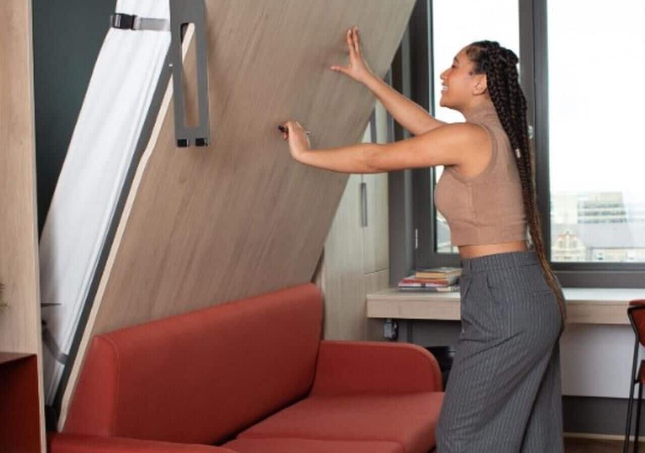 Iemand stelt een wandplank bij in een moderne kamer met een rode bank en een bureau.