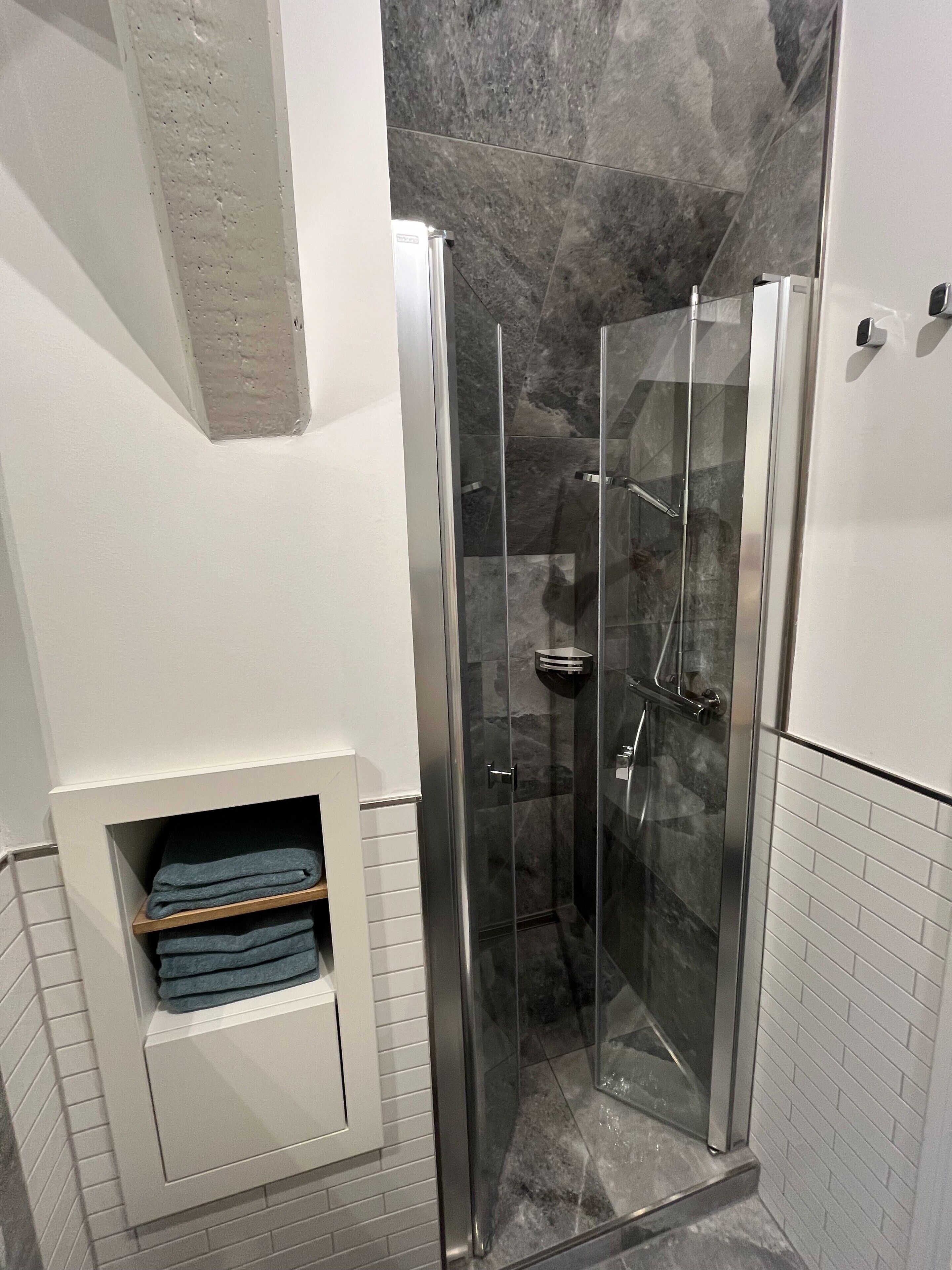 Un baño moderno con ducha envuelta en vidrio, paredes con azulejos blancos y un estante con toallas dobladas.