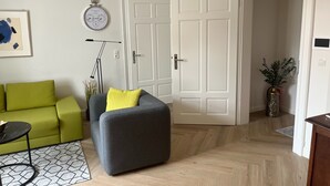 Un salon avec un canapé vert, un fauteuil gris agrémenté d’un coussin jaune, une lampe sur pied, une horloge et une porte donnant sur une autre pièce.