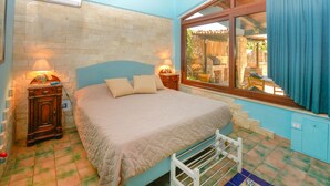 Une chambre à coucher avec un lit, deux tables de chevet, une fenêtre avec des rideaux, un mur en pierre et un sol carrelé.