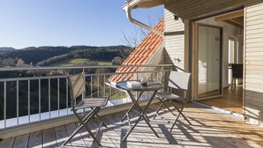Ein Balkon mit Blick auf ein bewaldetes Tal, ein kleiner Tisch mit Stühlen und eine Schiebetür zu einem Innenraum.