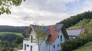 A modern house with a sloped roof, solar panels, and a balcony overlooking a hilly landscape. - Apartment 'Dollenberg' with Mountain View, Balcony and Wi-Fi (Bad Peterstal-Griesbach)