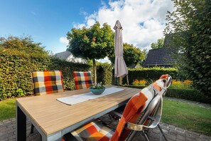 Un espace extérieur aménagé pour les repas, avec une table en bois, des chaises dotées de coussins à motifs, un vase de fleurs et un grand parasol.