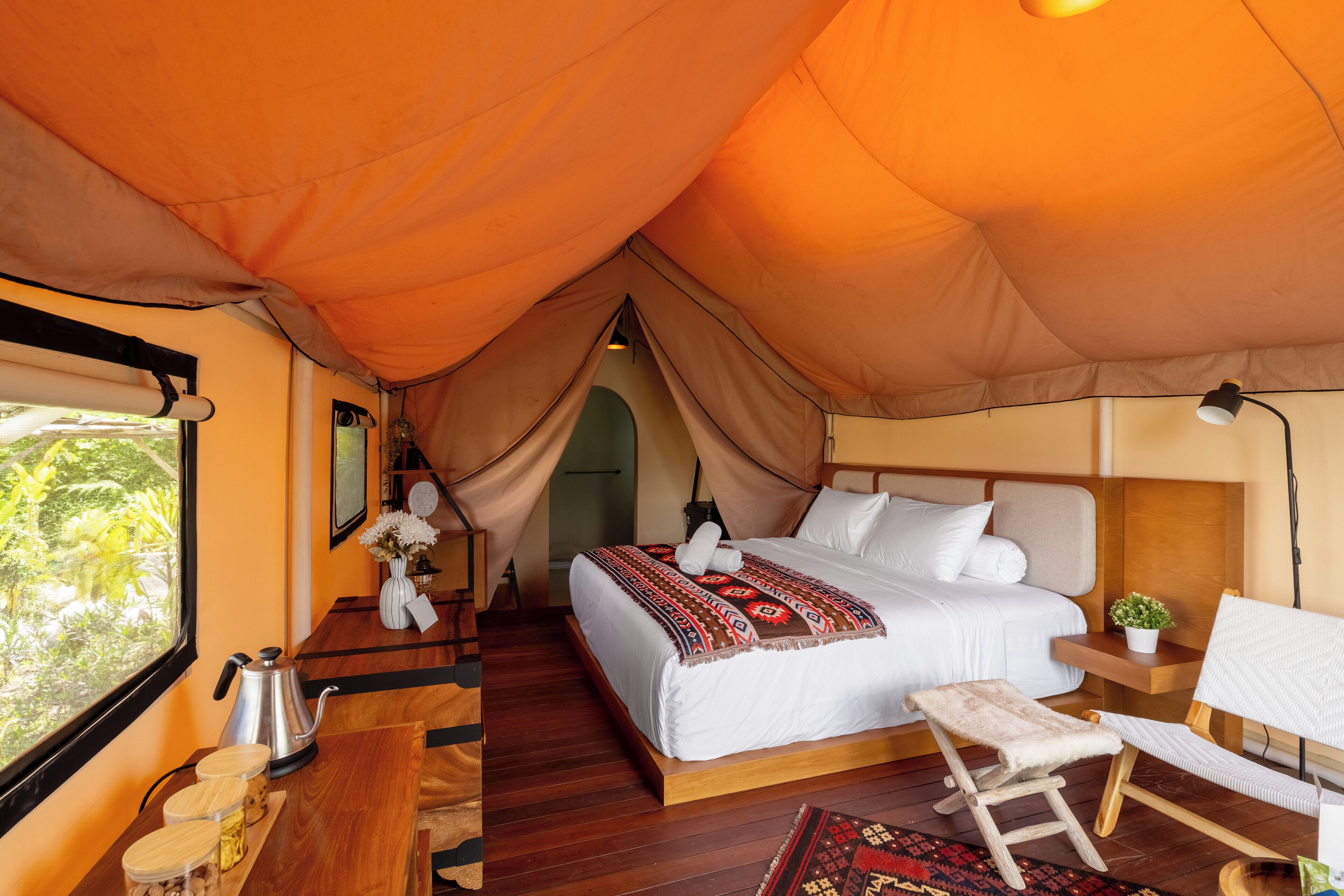 A tented room with a bed, bedside table, chair, and a view of greenery outside.