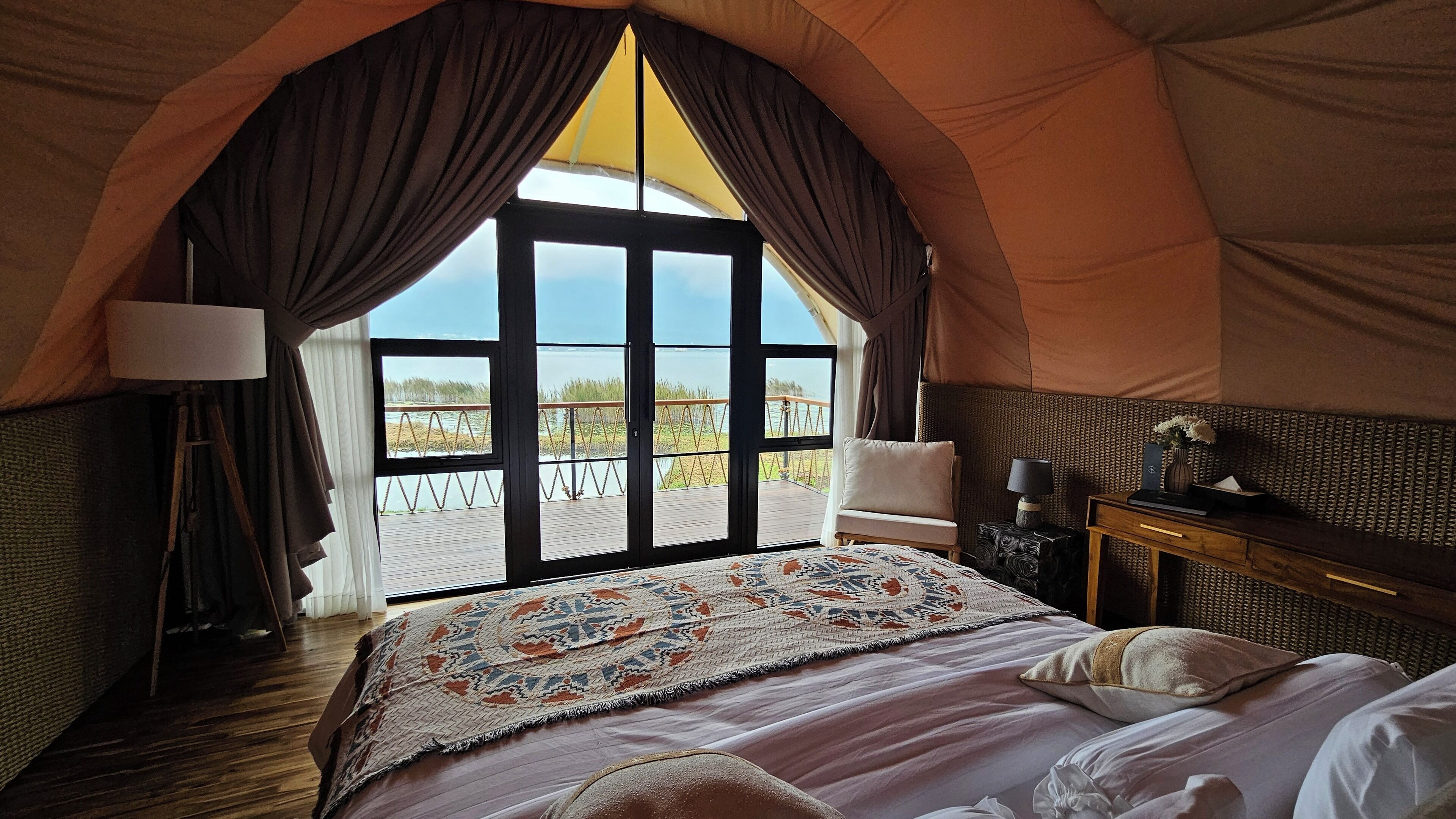 A tented room with a large bed, a desk, a lamp, and a view of the beach.