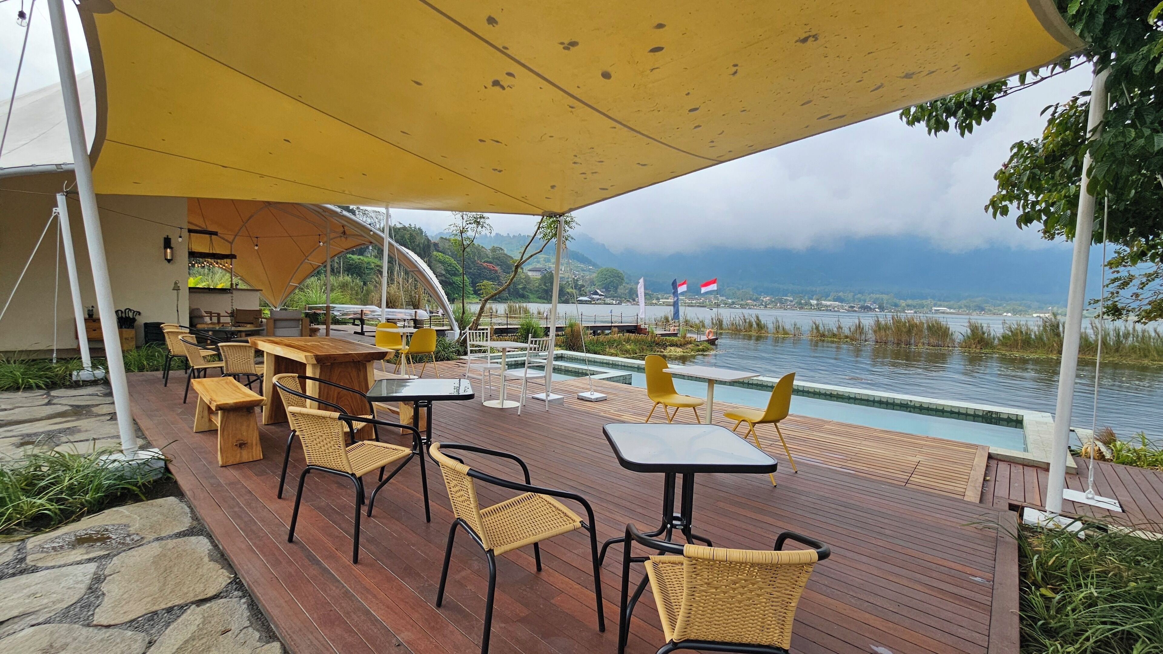 Een buitenterras met houten tafels en stoelen, een zwembad en uitzicht op een meer en bergen.