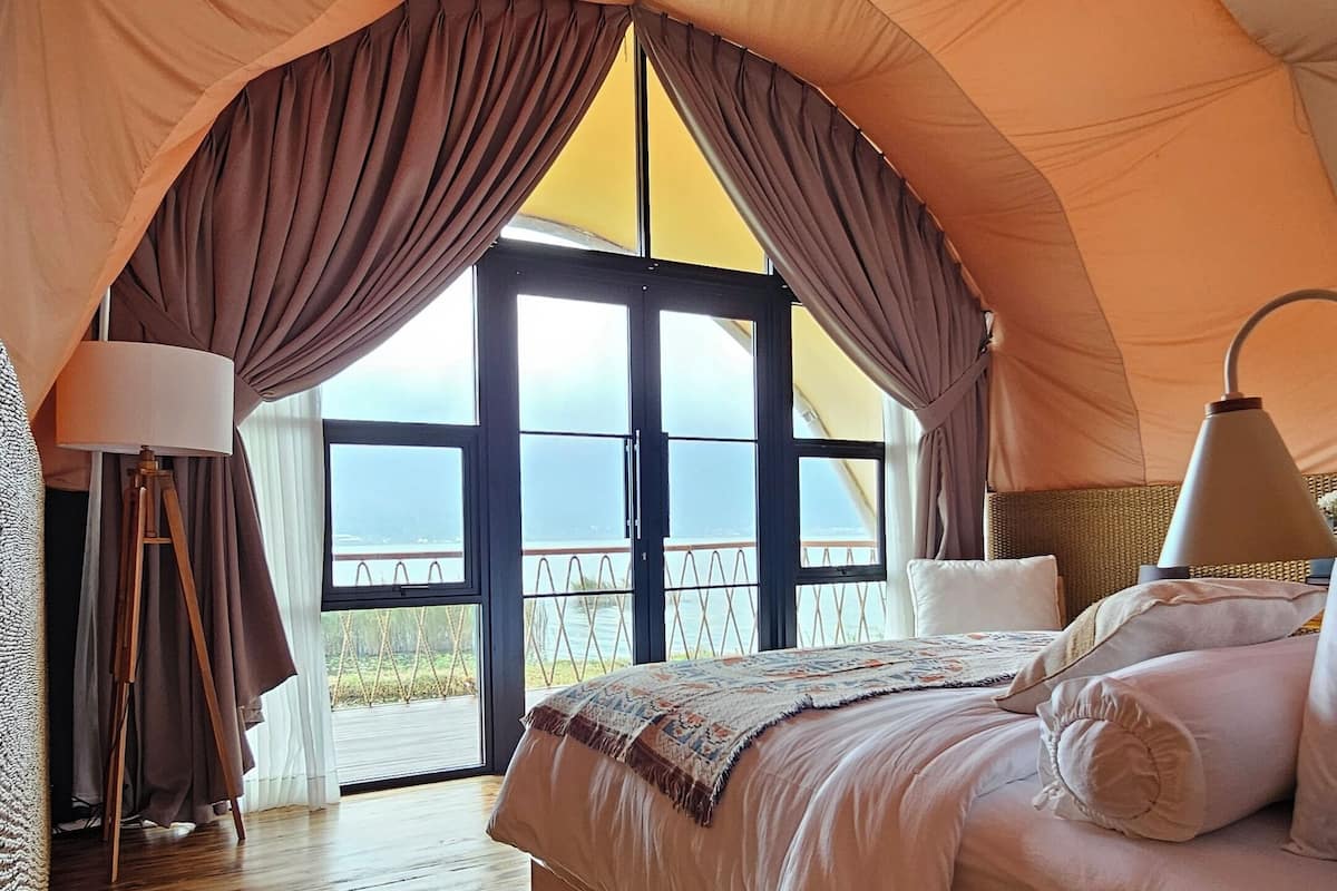 A tented accommodation room with a bed, a lamp, and a view of the sea through large windows.