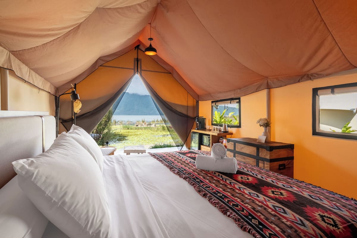 A tented room with a bed, a small table, a lamp, and a view of the outdoors.