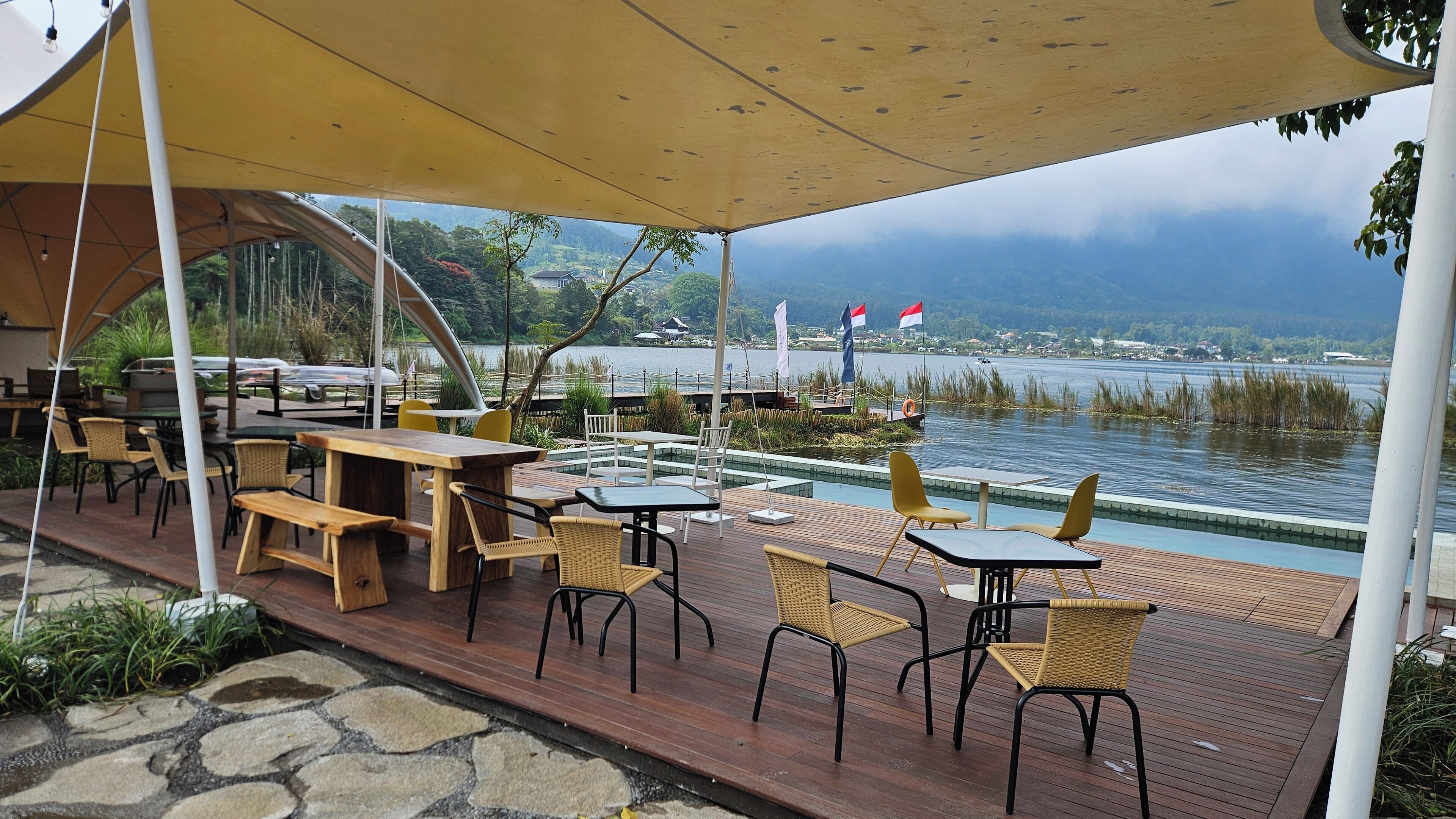 Un espace extérieur aménagé pour les repas, avec des tables et des chaises, une terrasse en bois et une vue sur un lac où flottent des drapeaux.