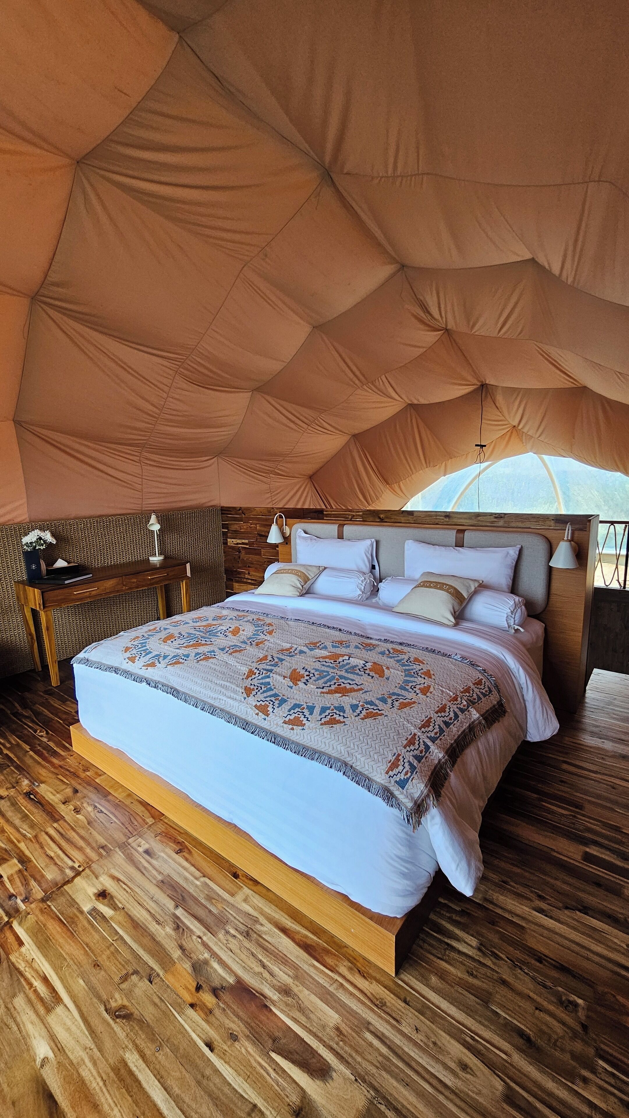 Una carpa espaciosa con una cama grande, suelo de madera y un escritorio con lámpara.
