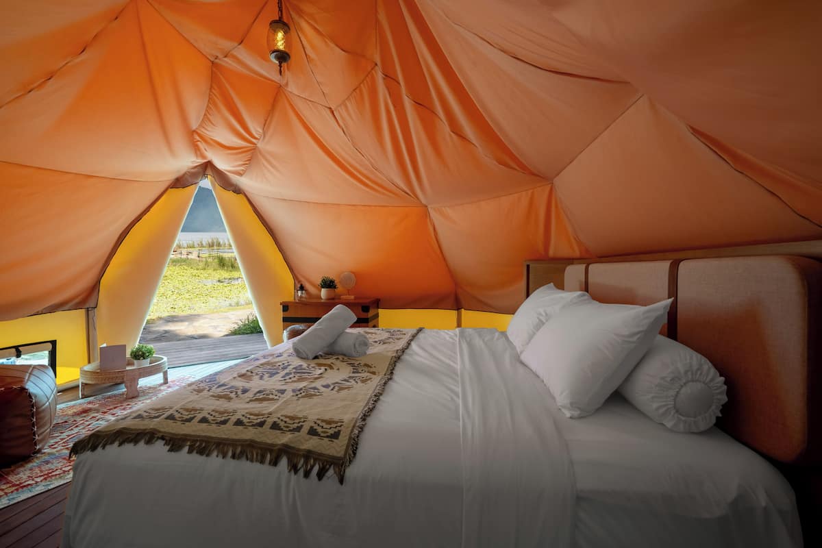A tented accommodation with a bed, pillows, a blanket, and a nightstand with a lamp.
