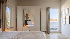 Ein Schlafzimmer mit einem großen Bett, einem Kleiderschrank, einem Spiegel und Blick ins Freie durch ein Fenster.