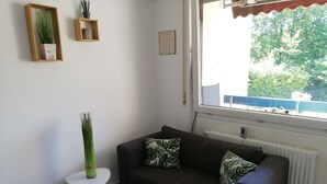A living room with a dark brown sofa, two green patterned pillows, a white side table with a tall green vase, and a window with a view of trees and a blue railing. - Apartment 'Lived-in' with Mountain View, Balcony and Wi-Fi (Bad Urach)