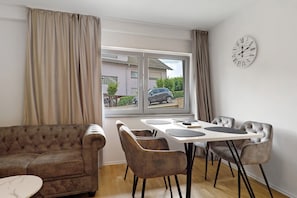 Et værelse med sofa, spisebord, stole, et ur og et vindue med udsigt til en bygning og en bil.