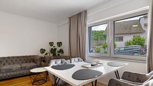 A modern living room with a sofa, a dining table with chairs, a potted plant, and a view of a residential area through large windows. - Apartment 'Ferienwohnung' with Private Terrace, Private Garden and Wi-Fi (Sinsheim)