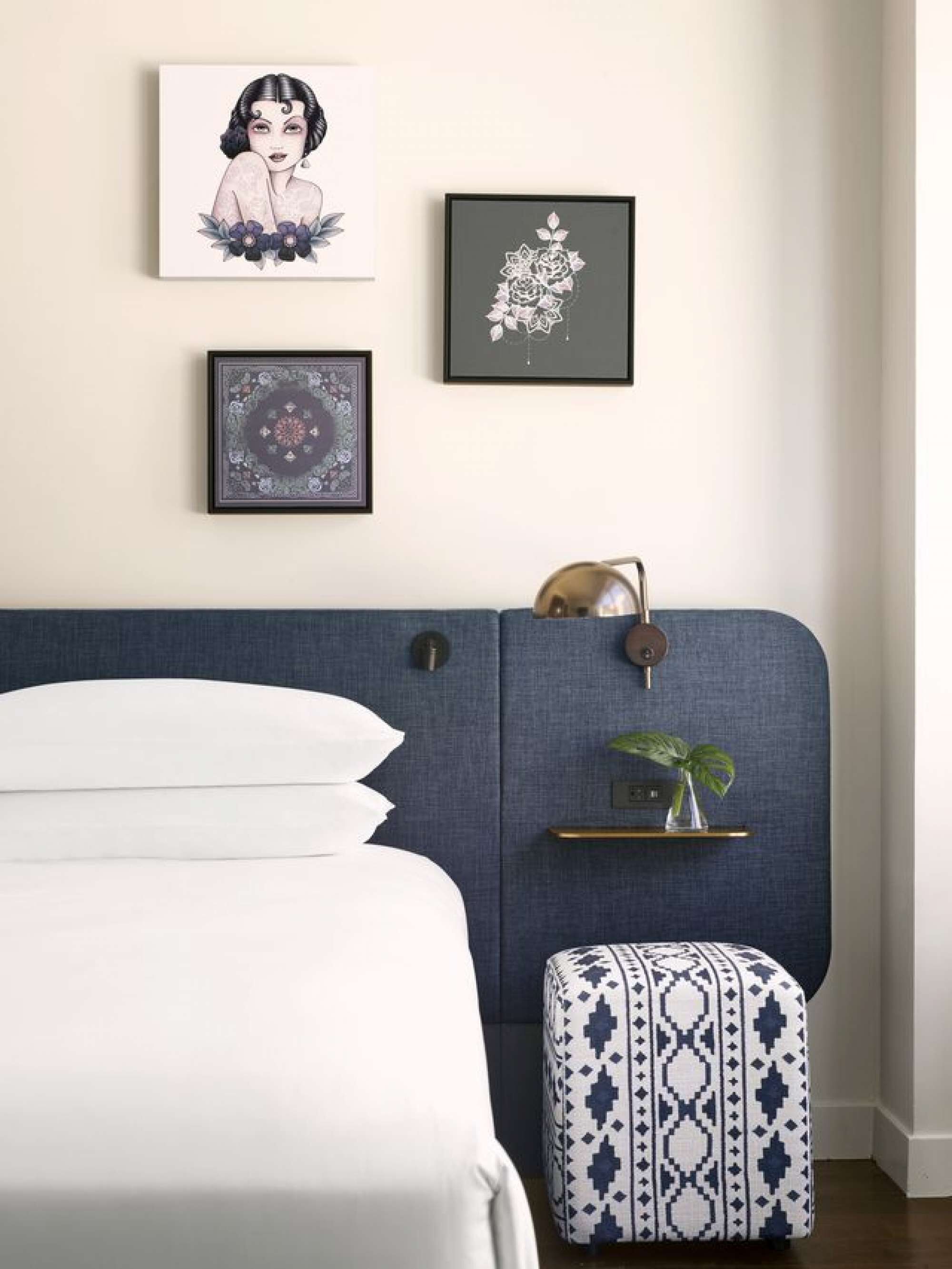 A neatly arranged bedroom with a bed, a bedside table with a plant, and three framed artworks on the wall.