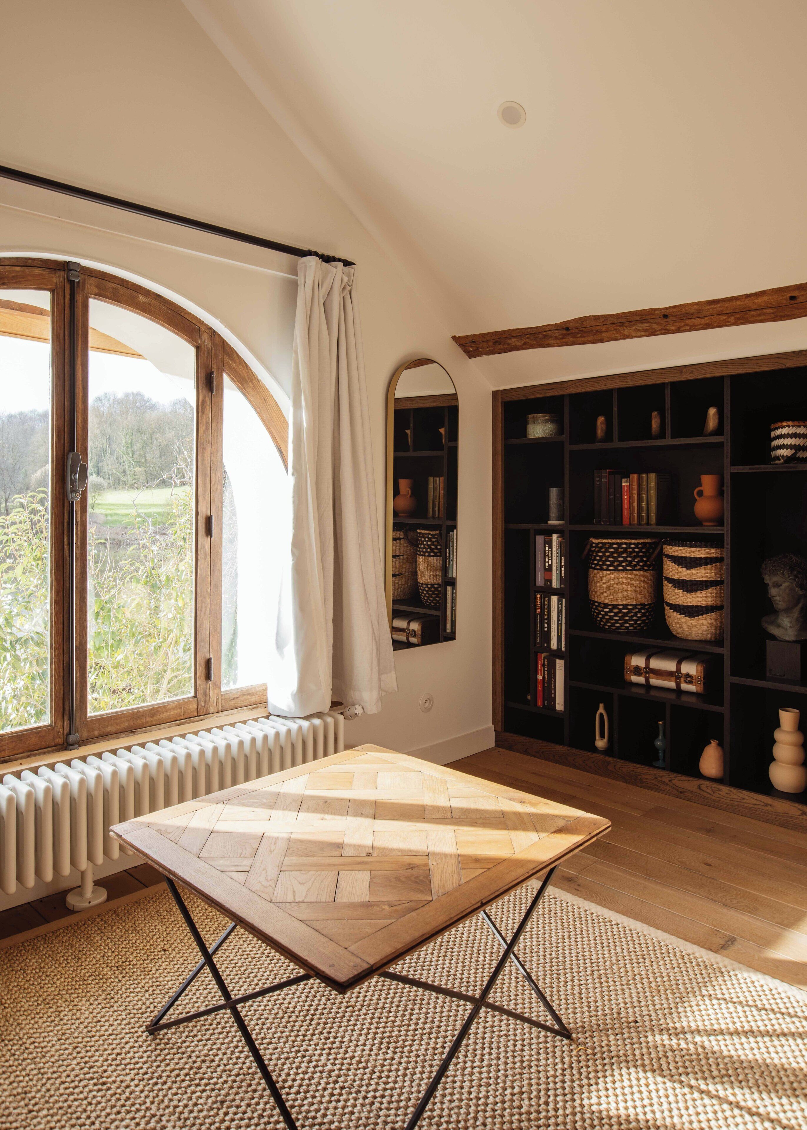Ein Zimmer mit großem Fenster, einem Holztisch, einem Bücherregal und einer Heizung.