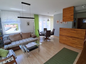 A modern living room with a sofa, a glass coffee table, a dining table with chairs, and a kitchen area with wooden cabinets.