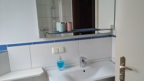 Un baño con un lavamanos blanco, un espejo encima y una ducha a la vista al fondo.