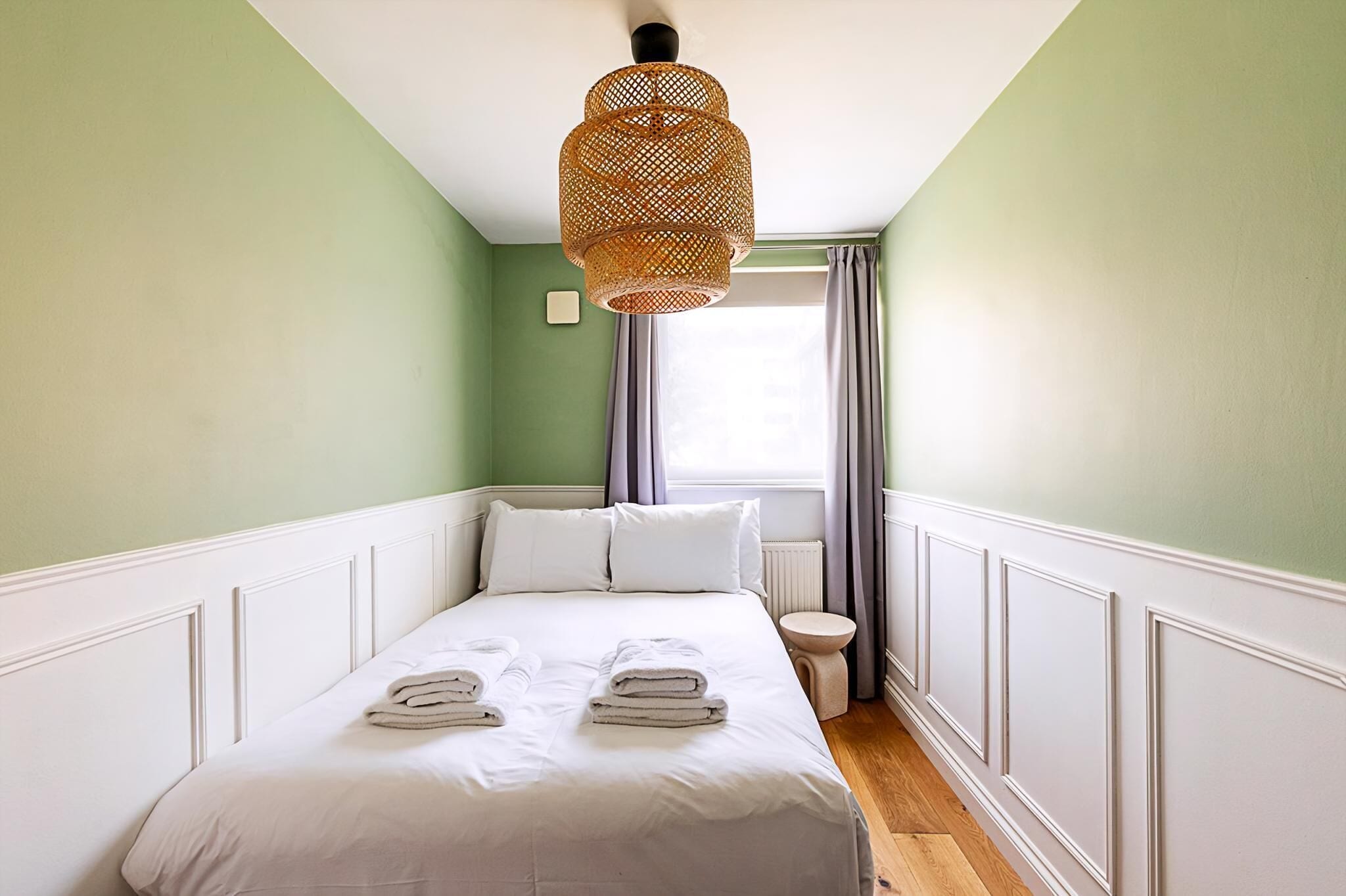 A neatly made bed with white linens and towels, a wicker hanging light fixture, a window with curtains, and a radiator.