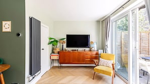 A modern living room with a wooden cabinet, a television, a yellow armchair, and a large window. - Stylish Islington Garden Flat Sleeps Four (London)