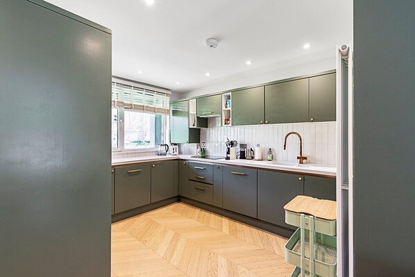 A modern kitchen with grey cabinets, a wooden herringbone floor, and a window with blinds. - Stylish Islington Garden Flat Sleeps Four (London)