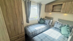 A compact bedroom with two beds, a wardrobe, and a window with curtains. - Smart, modern two bedroom caravan, perfect for couples and families alike. (Aldbrough)