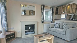 A living room with a fireplace, a sofa, a coffee table, and wooden cabinets. - Smart, modern two bedroom caravan, perfect for couples and families alike. (Aldbrough)