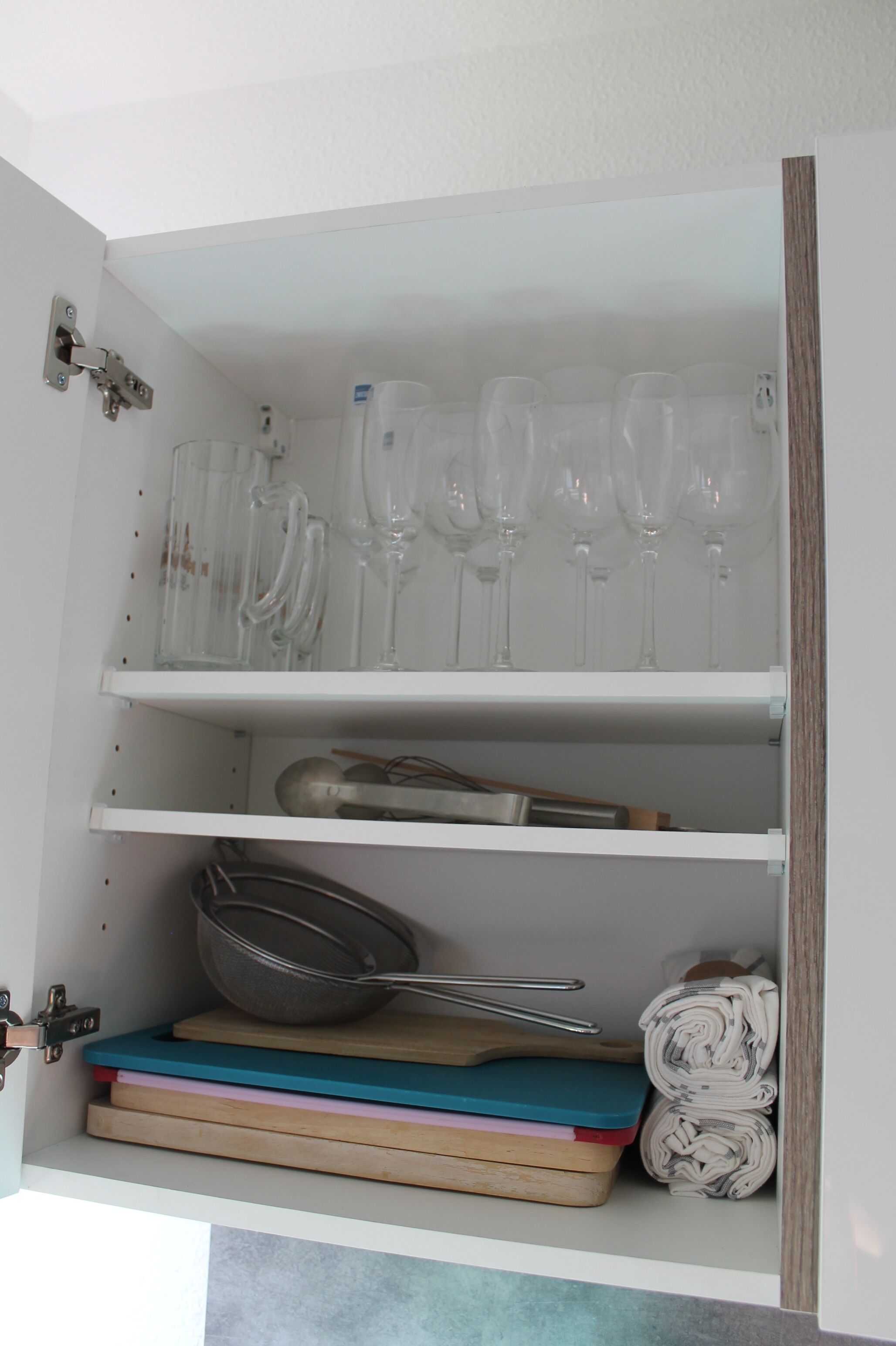 A cabinet with shelves containing wine glasses, a strainer, and rolled newspapers.