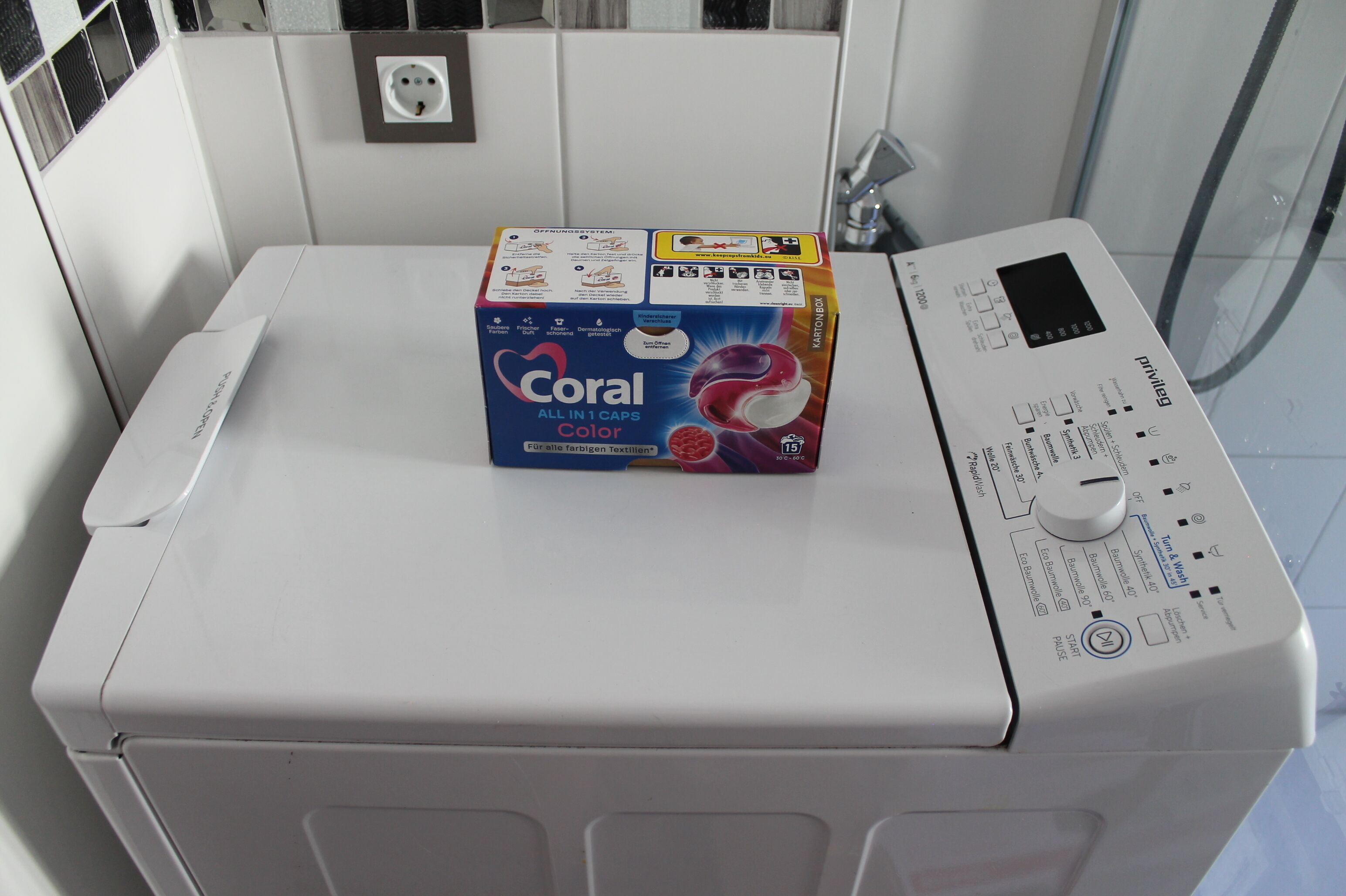 Eine Waschmaschine mit Frontlader, ein Bedienfeld und eine Packung 'Coral All in 1 Caps Color' Waschmittel darauf.