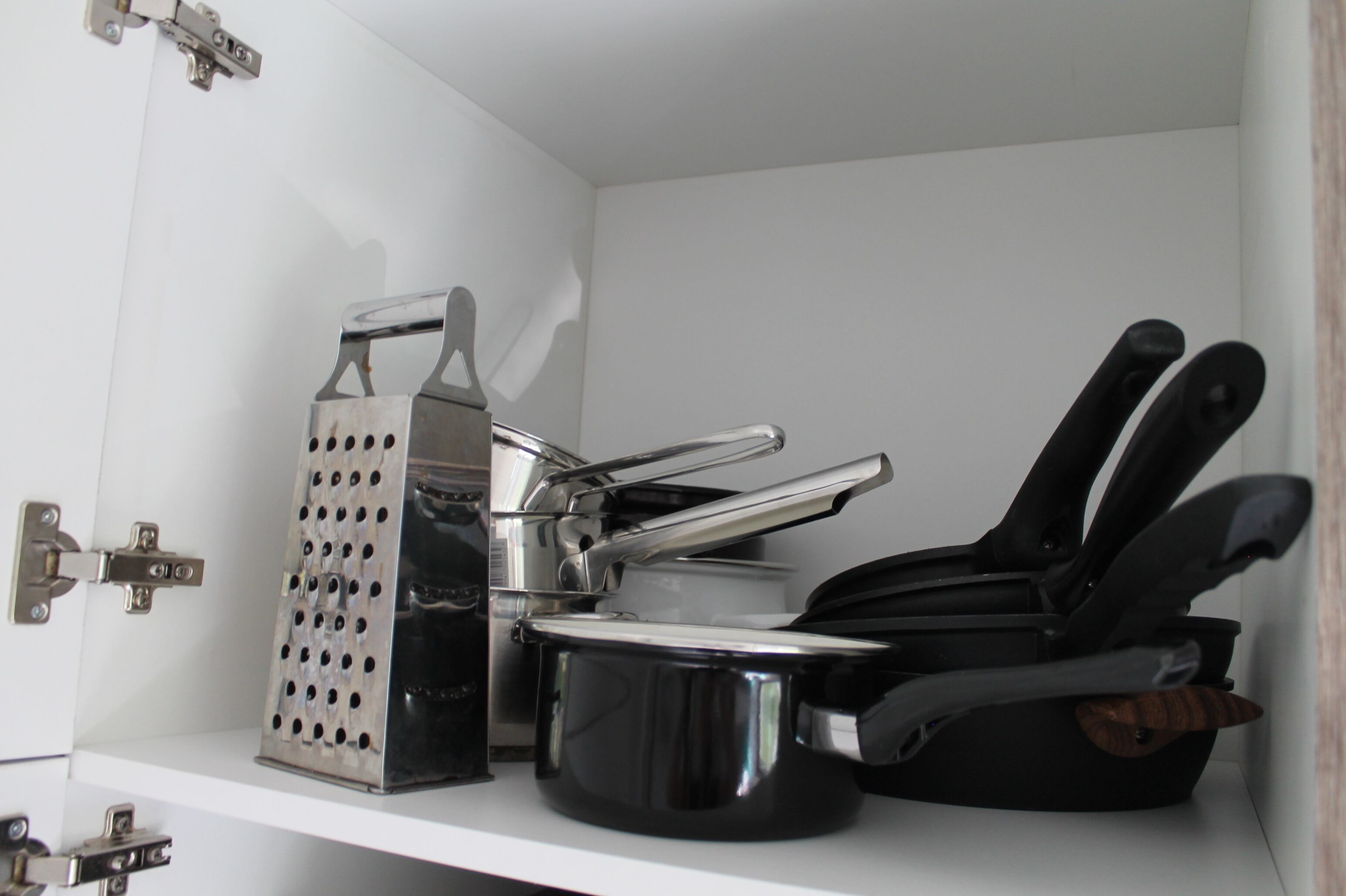 Une cuisine avec un meuble blanc, une casserole noire, une râpe et un ensemble de couteaux noirs.