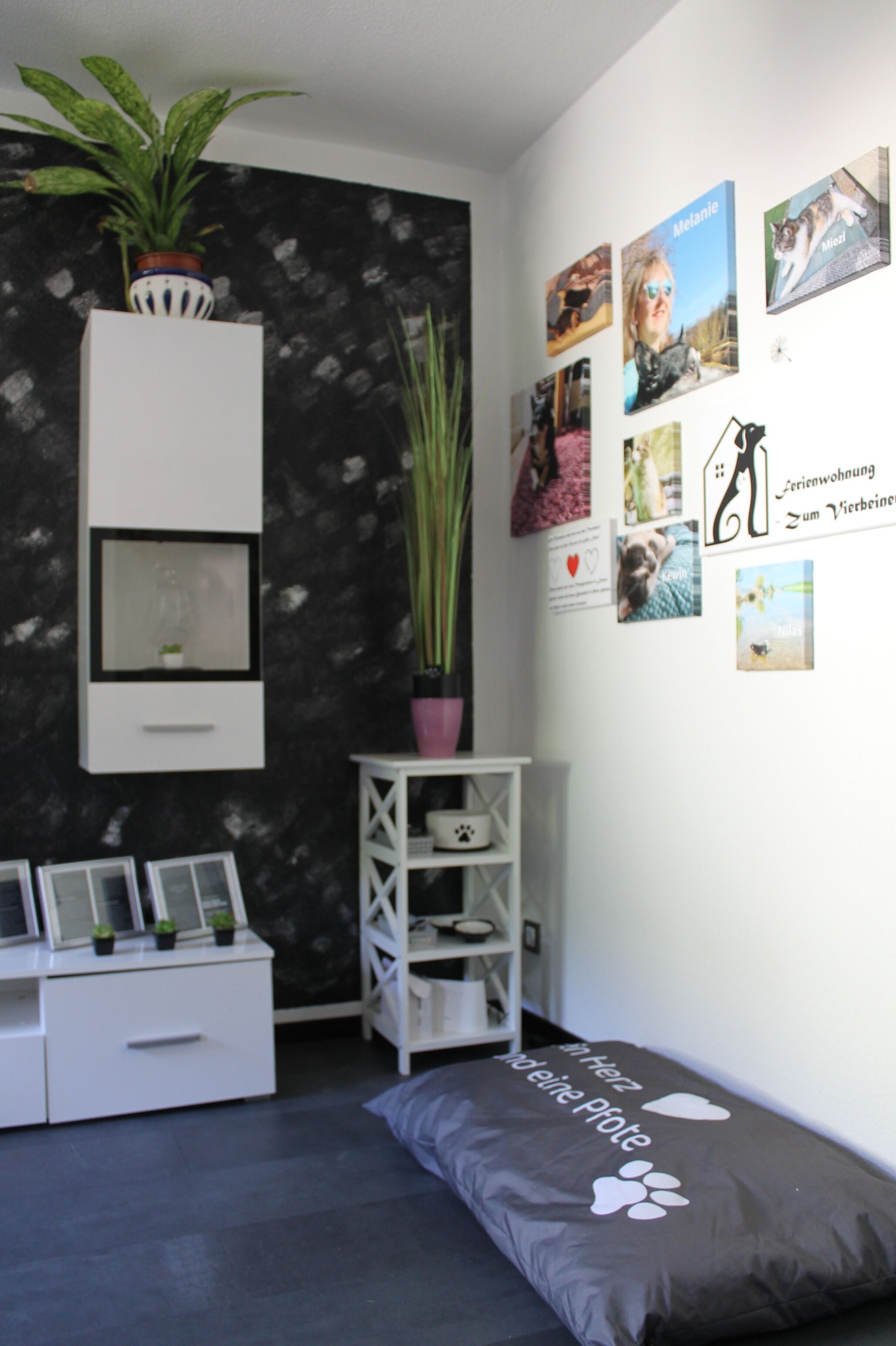 Ein Schlafzimmer mit einem Bett, einem Nachttisch, einer kleinen Ablage, einer Topfpflanze und einer Wand mit Bildern und Text.