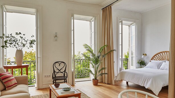 Habitación amplia con cama, sofá, mesa de comedor y una silla. Tiene un balcón con plantas y vistas a zonas verdes.