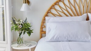 Una cama con ropa blanca, un cabecero redondo tejido, una mesita de noche pequeña blanca con un florero y una lámpara fijada a la pared.