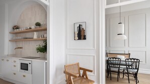 Eine moderne Küche mit weißen Schränken, einem eingebauten Backofen und einem Holzregal. Es gibt einen Essbereich mit Tisch, Stühlen und einer Pendelleuchte.
