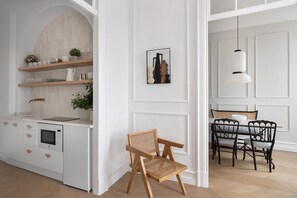 A modern kitchen with white cabinetry, a built-in oven, and a wooden shelf. There is a dining area with a table, chairs, and a hanging light fixture. - Suite By Olivo (Vigo)