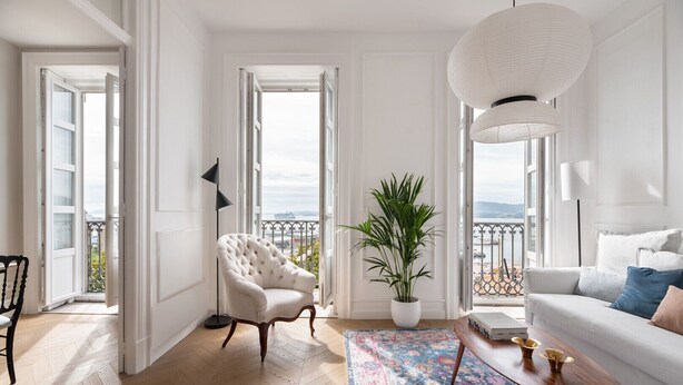Ein geräumiges Wohnzimmer mit einem weißen Sofa, einem gesteppten Sessel, einer Topfpflanze und einem großen Fenster mit Blick auf das Meer.