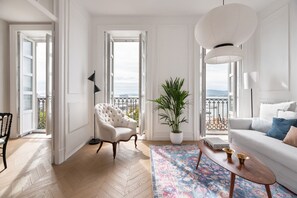 A spacious living room with a white sofa, a tufted armchair, a potted plant, and a large window offering a view of the sea. - Suite By Olivo (Vigo)