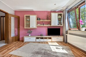 Una sala de estar moderna con pared rosa, piso de madera, televisor empotrado y un ventanal con vistas a árboles.