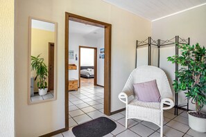 Ein Flur mit einem weißen Korbstuhl, einem Blumentopf und einem Spiegel, der einen Raum mit Bett und Kommode widerspiegelt.